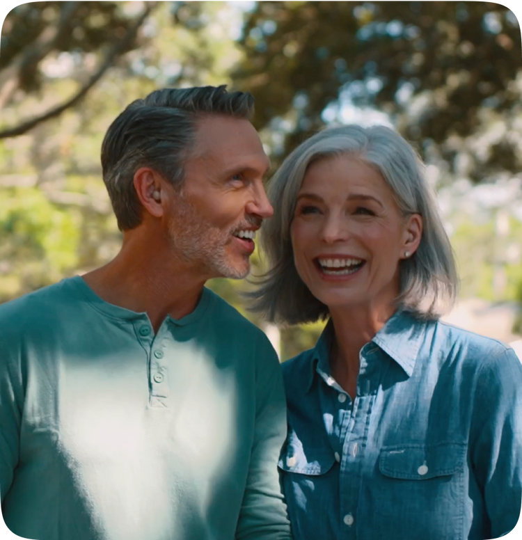 Man and woman smiling at each other outdoors with trees in the background

